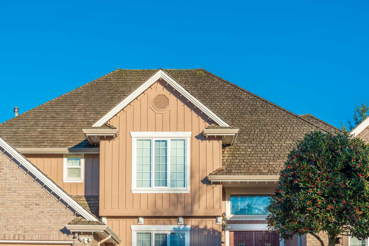 The exterior of a two story house is seen on a clear day. The house has beige siding and a brown roof. A large green leafy tree with red berries partially blocks the front door. The sky is bright blue.